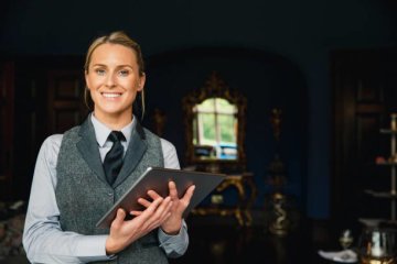 A shot of a beautiful blonde hotel receptionist standing inside a hotel, holding a digital tablet and looking positive.